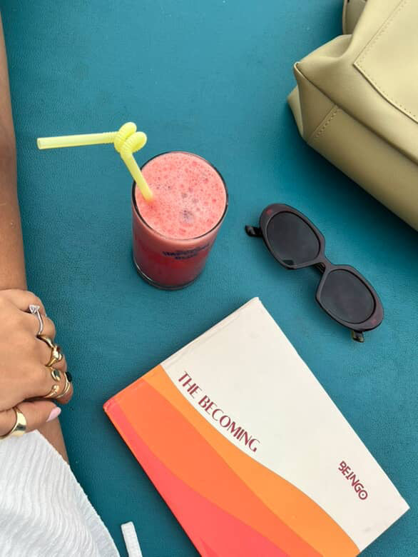 Smoothie with a straw, sunglasses, and a book titled &#39;The Becoming&#39; on a teal surface.